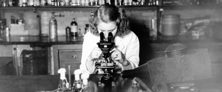 A black and white photo of a woman looking into a microscope in 1948